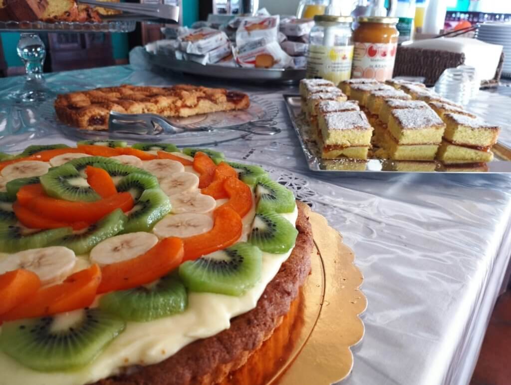 Tavola imbandita con dolci assortiti, tra cui una torta alla frutta con kiwi, arance e banane.